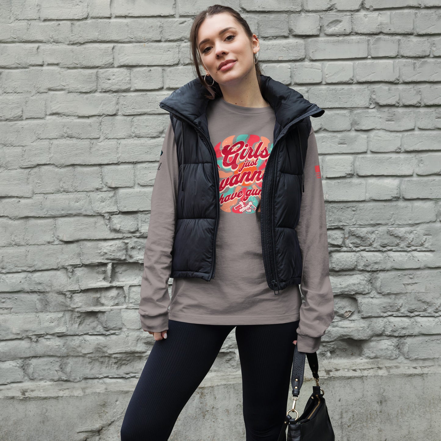 Woman wearing a gray long sleeve shirt with a black vest over a graphic t-shirt against a brick wall.