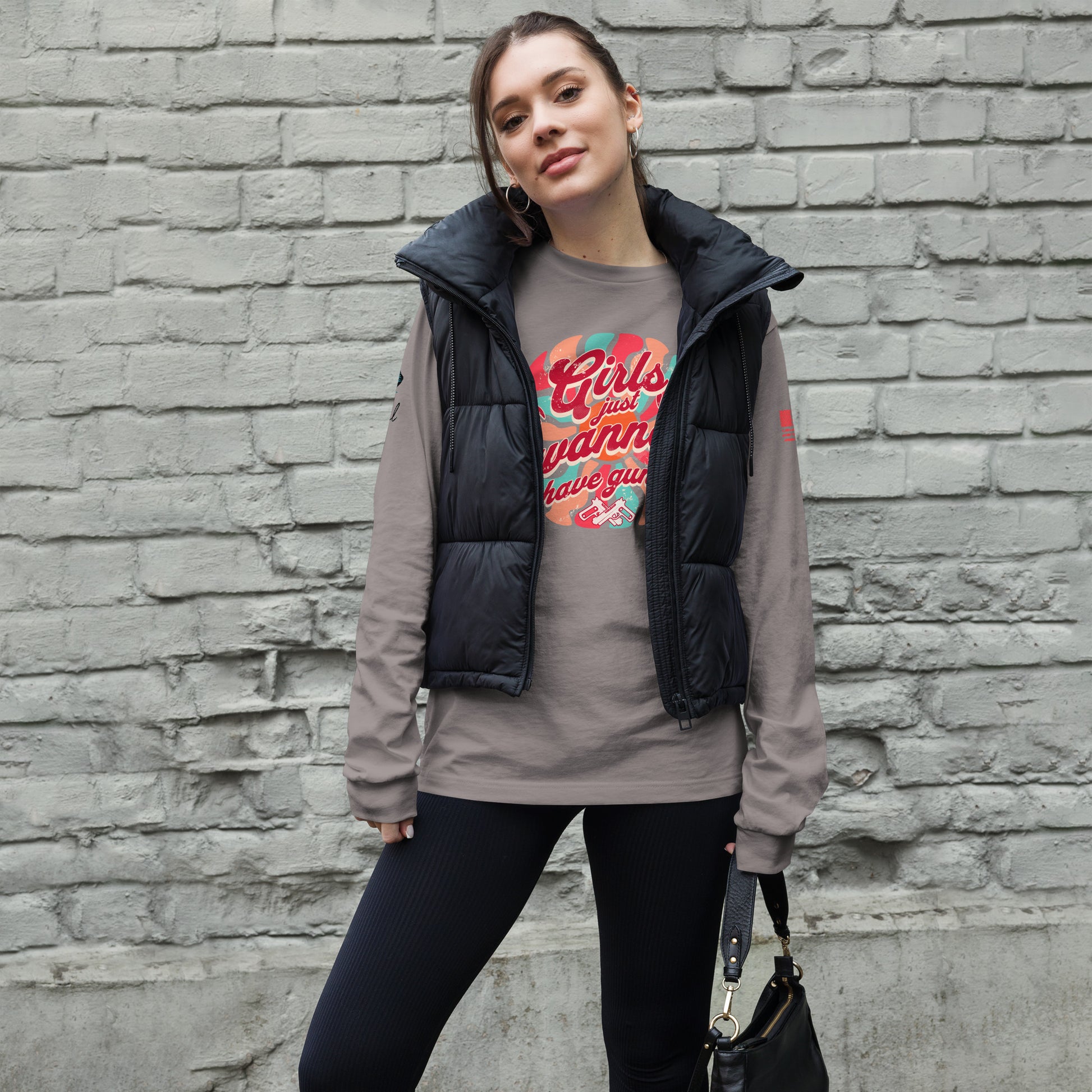 Woman wearing a gray long sleeve shirt with a black vest over a graphic t-shirt against a brick wall.