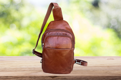 Brown leather sling bag on a wooden surface with a blurred green background