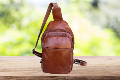 Brown leather sling bag on a wooden surface with a blurred green background
