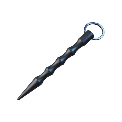 A tactical black training weapon, resembling a short bamboo staff with a split ring, placed diagonally on a textured gray stone tile with dark wood borders.